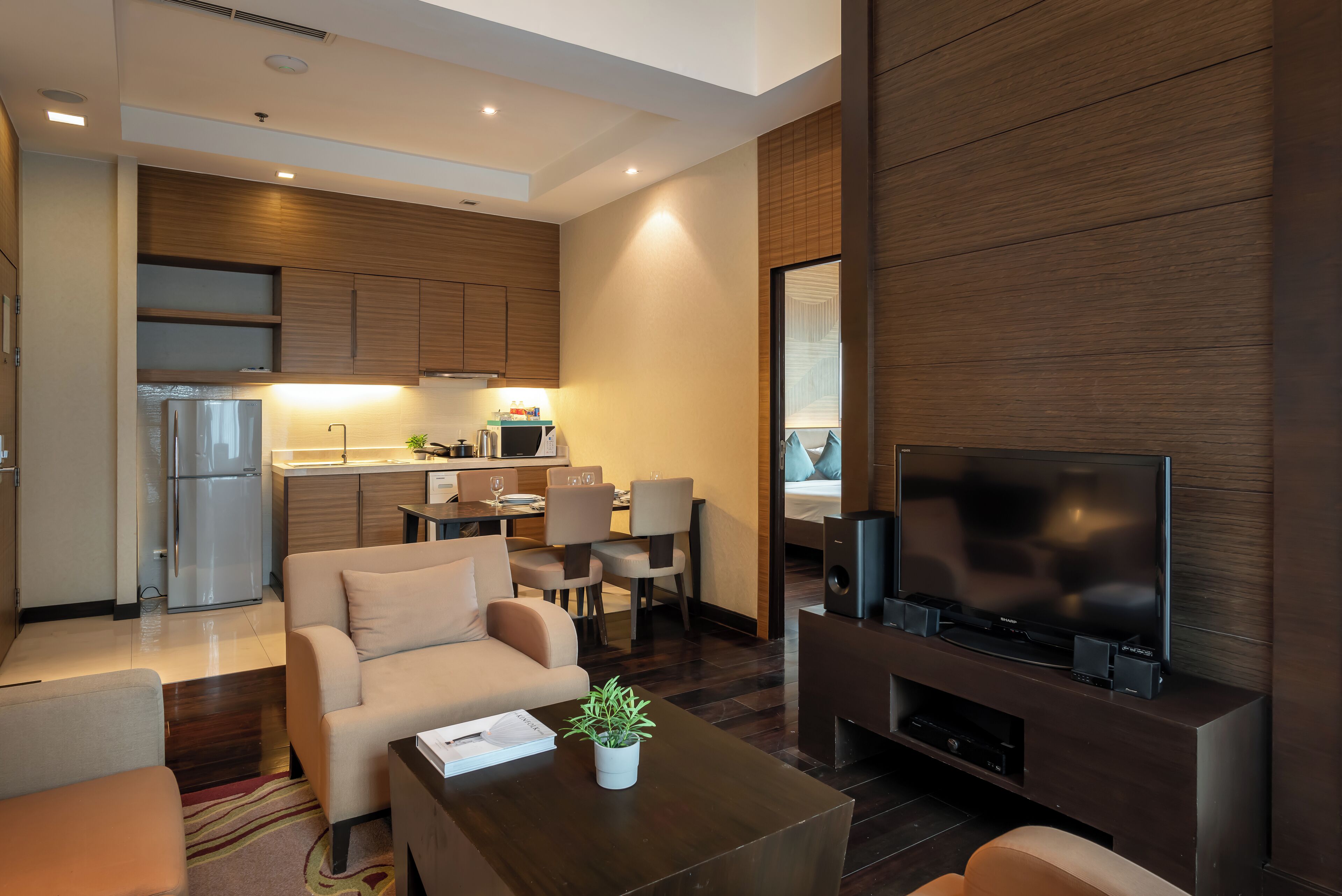A modern living room with a sofa, coffee table, and a TV. There is a kitchen area with a refrigerator and microwave in the background.