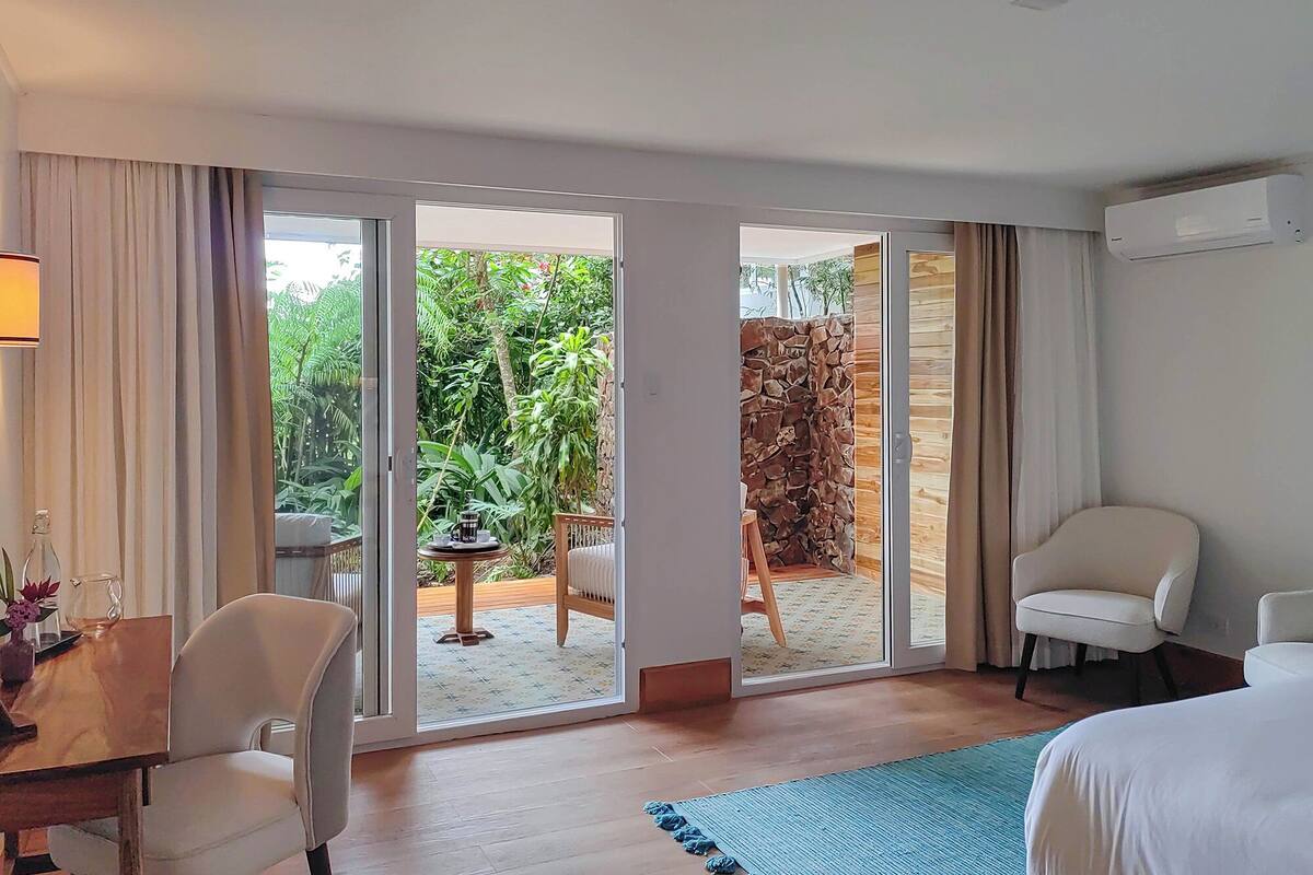 Ein Hotelzimmer mit einem Bett, zwei weißen Stühlen, einem kleinen Tisch mit einer Lampe und Blick auf einen Garten durch eine Schiebetür.