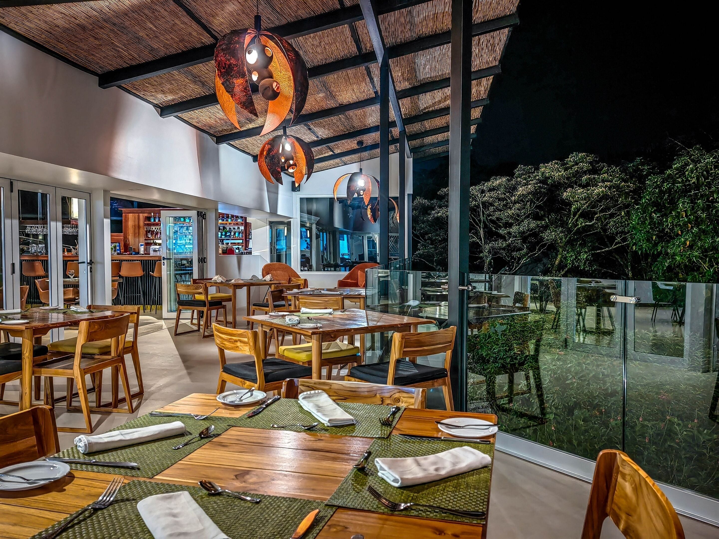 Un área de comedor al aire libre con mesas y sillas de madera, una barra con estantes llenos de botellas y vistas a zonas verdes a través de grandes ventanales.