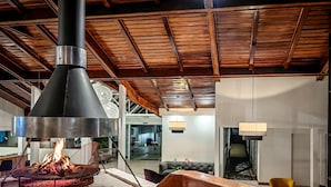 Un intérieur moderne avec un plafond orné de poutres en bois, un foyer central et un coin salon doté d’une cheminée en briques.