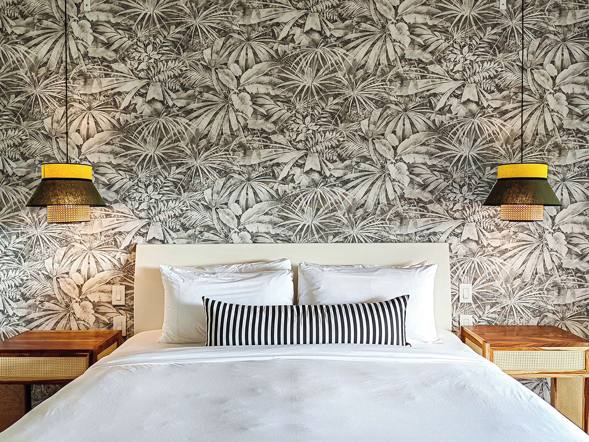 A bed with white bedding, two pillows, and a striped pillow, against a wall with a tropical leaf pattern. There are two nightstands with woven baskets and a hanging lamp with a yellow and black shade.