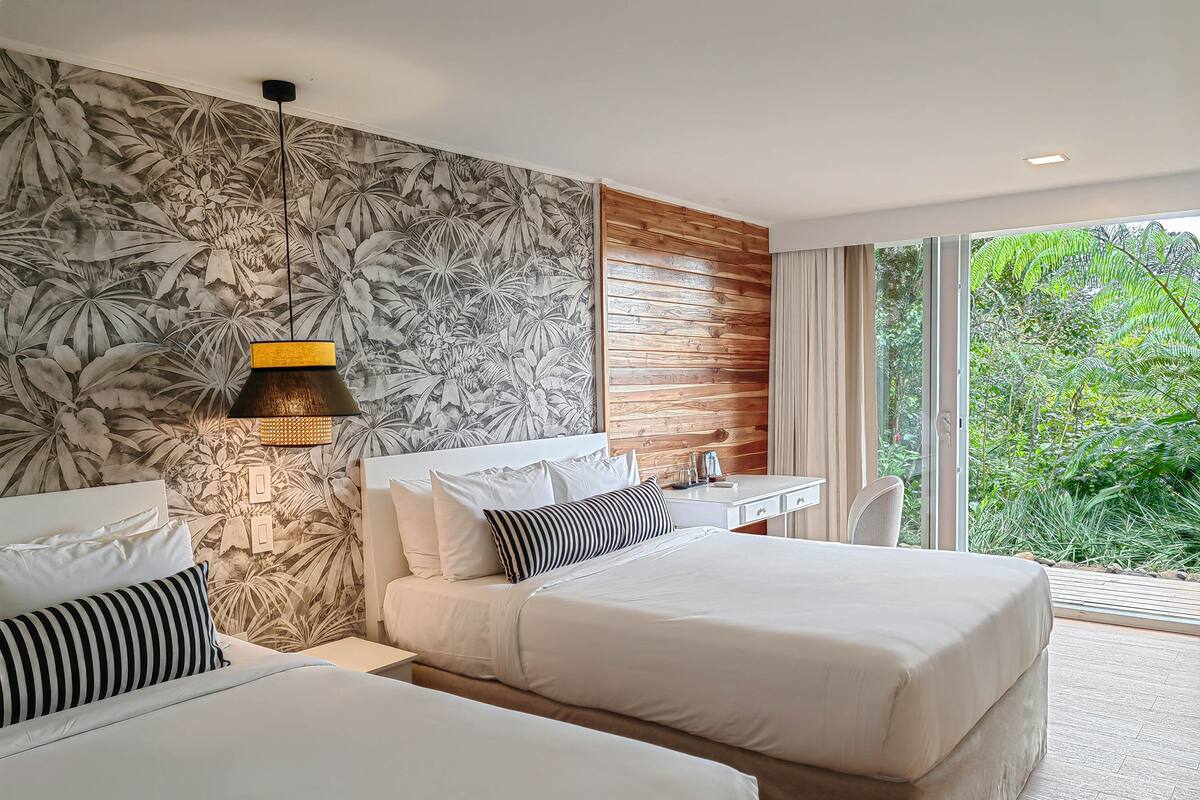 Ein Hotelzimmer mit zwei Betten, einem Schreibtisch und einem großen Fenster mit Blick auf Grünflächen.