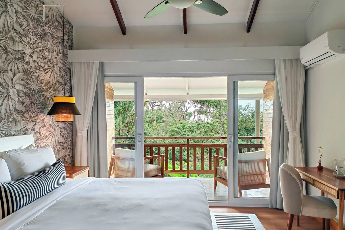 Ein Schlafzimmer mit Bett, Schreibtisch, Stuhl, Deckenventilator und einem Balkon mit Blick auf Grünflächen.