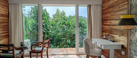 A modern hotel room with a large sliding glass door leading to a lush garden. There is a bed, a desk, and two chairs.