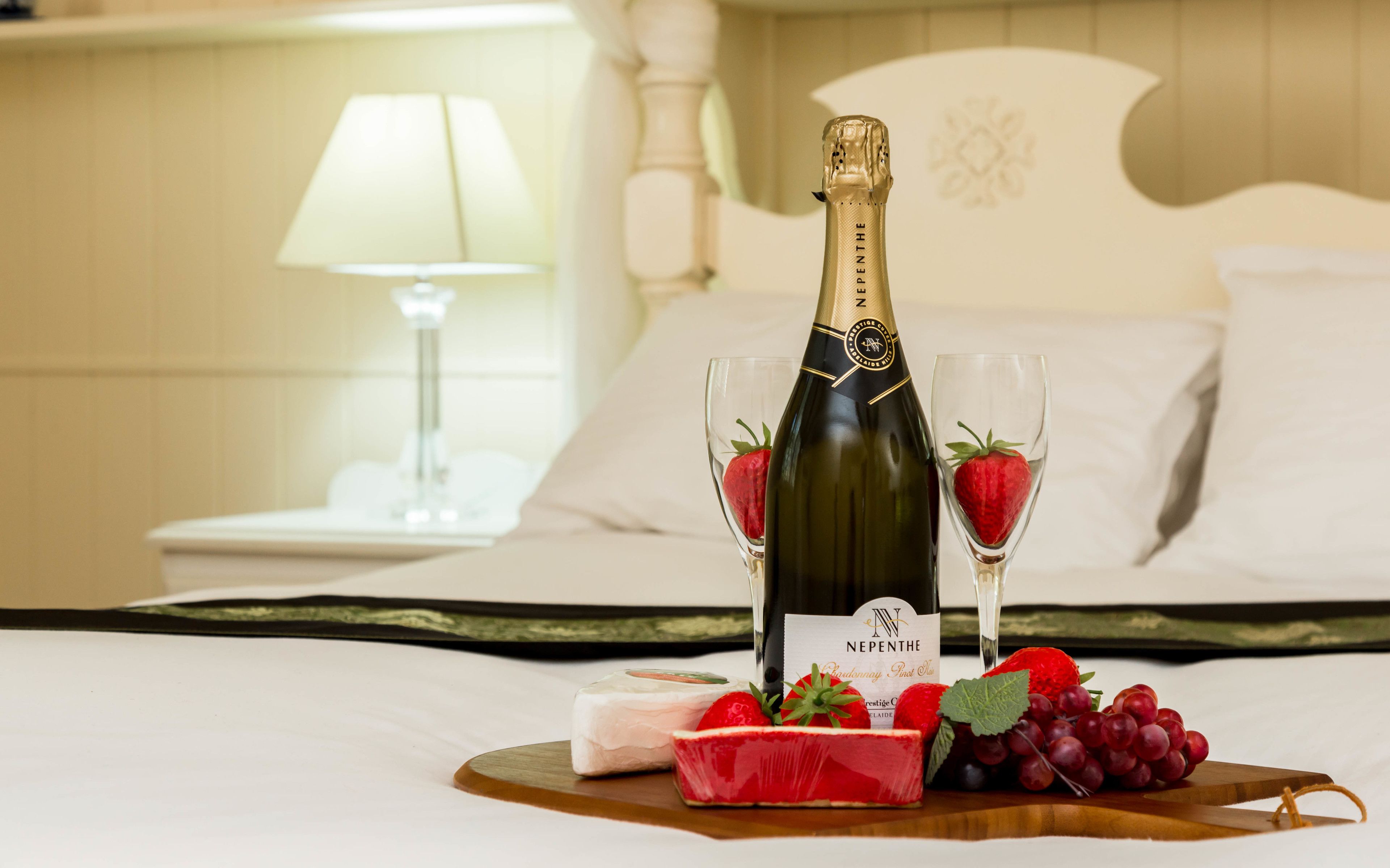 A bottle of Nepenthe champagne, two glasses with strawberries, a slice of red fruit cake, and grapes on a wooden board on a bed.
