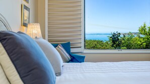 A bedroom with a large window offering a view of the sea and greenery.