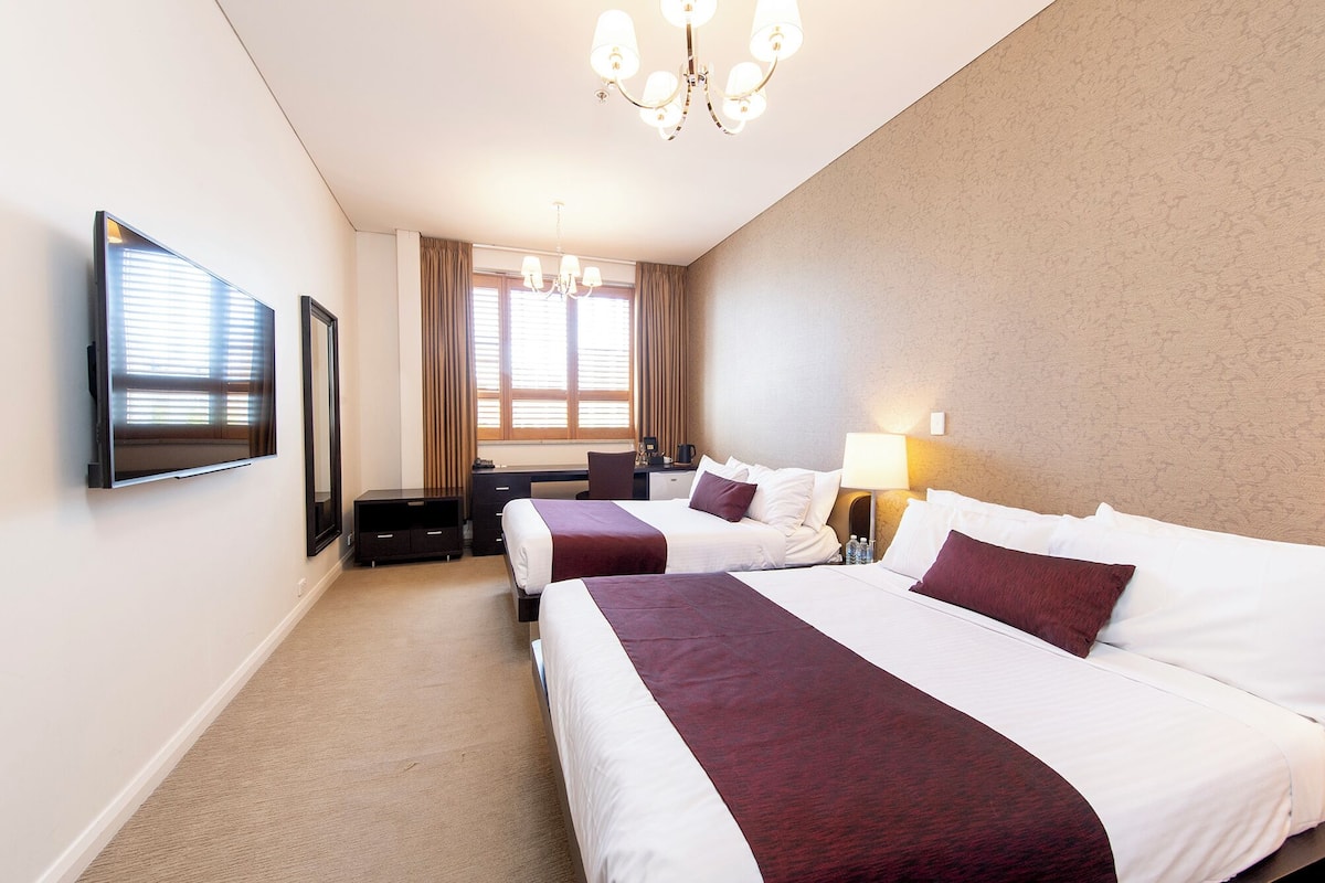 A hotel room with two beds, a desk, a chair, a TV, and a chandelier.
