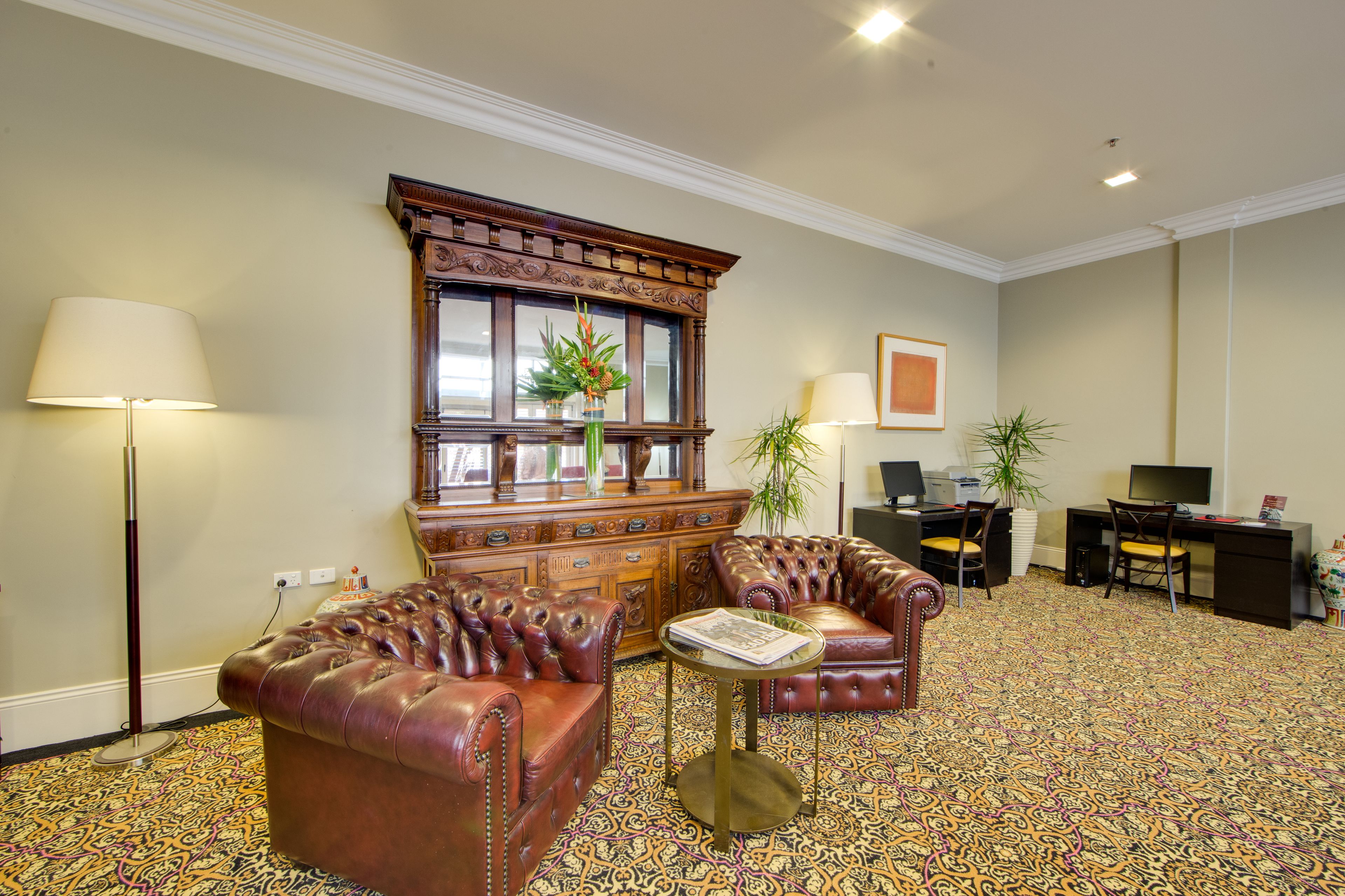 Una habitación con alfombra estampada, sillones de cuero, un mueble de madera, un escritorio con computadora y una lámpara de pie.
