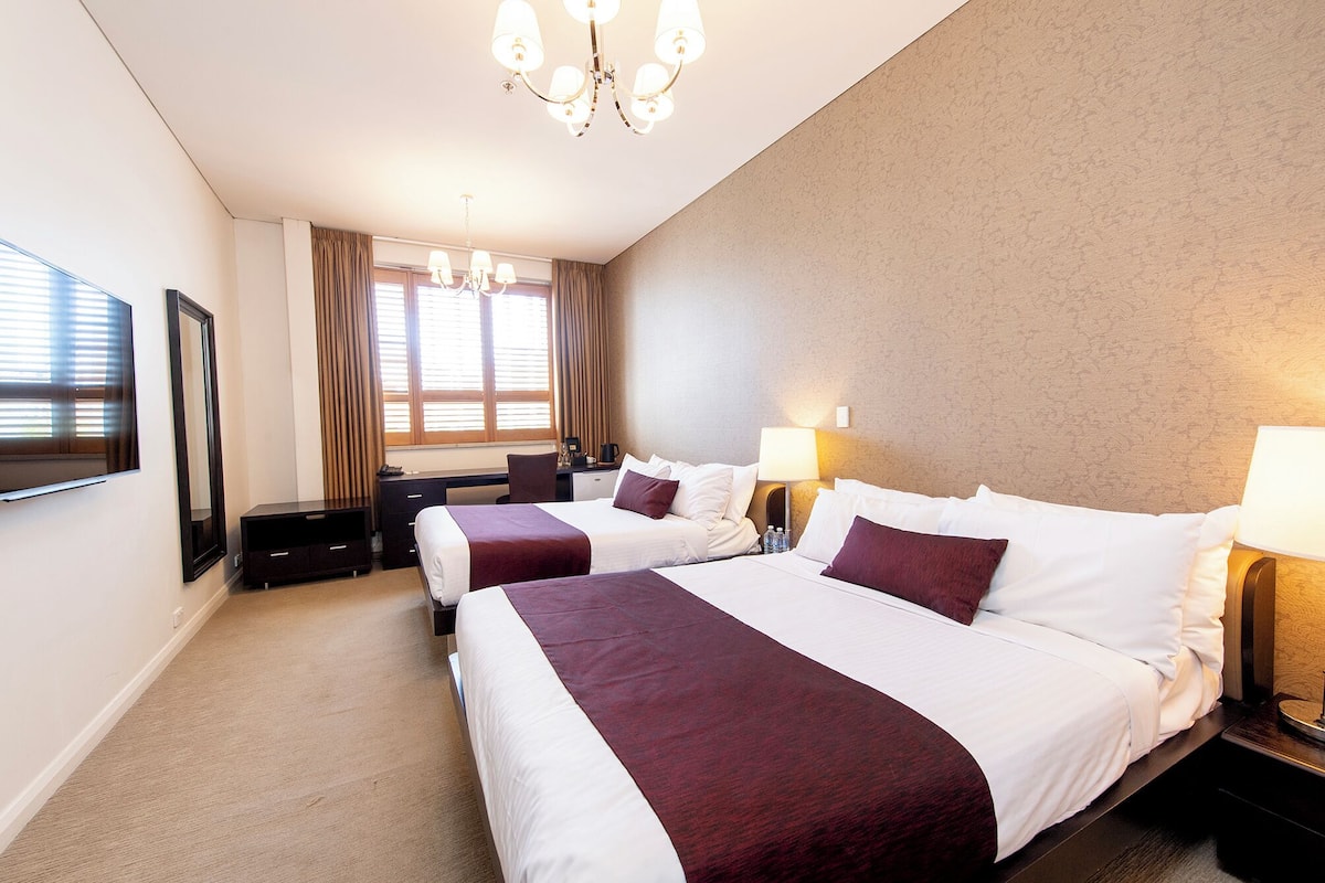 A hotel room with two beds, a desk, a chair, a TV, and a chandelier.