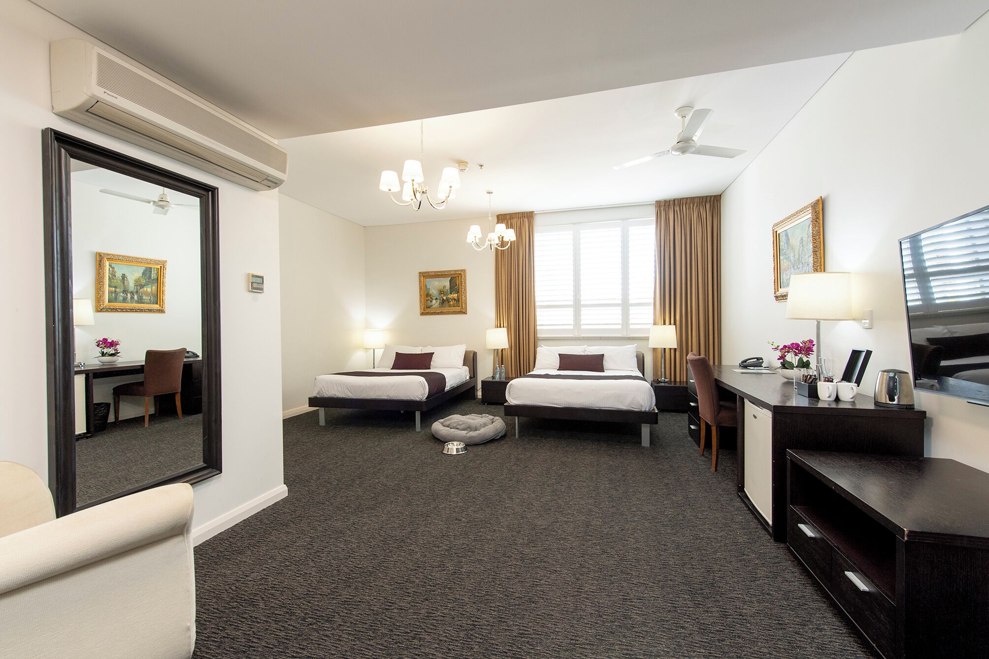 A hotel room with two beds, a desk, a chair, a desk lamp, a bedside table, a mirror, a ceiling fan, and a window with curtains.