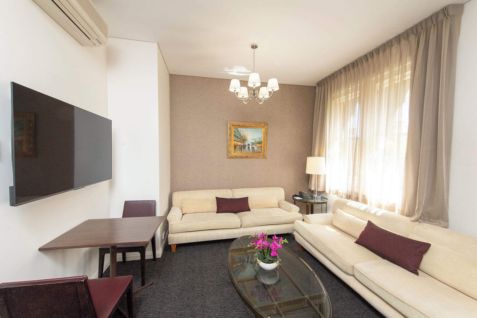 A hotel room with a flat-screen TV, a sofa, a coffee table with flowers, a painting on the wall, and a window with curtains.