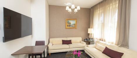 Habitación de hotel con televisión de pantalla plana, sofá, mesa de centro con flores, un cuadro en la pared y ventana con cortinas.