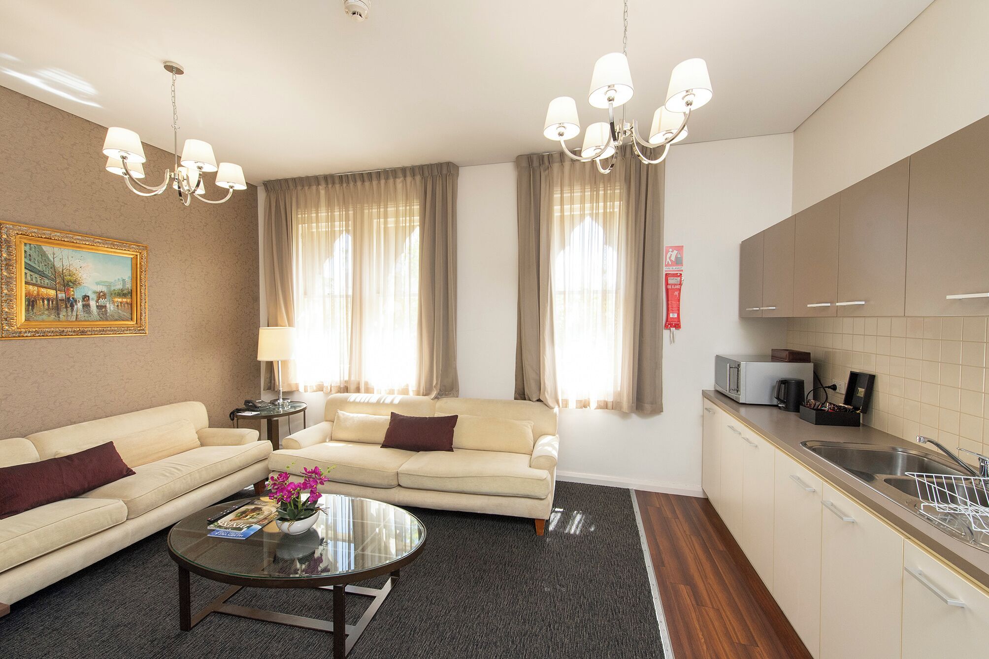 A modern hotel room with a sofa, a round coffee table with flowers, a dining area with a microwave, and a kitchen with a sink.