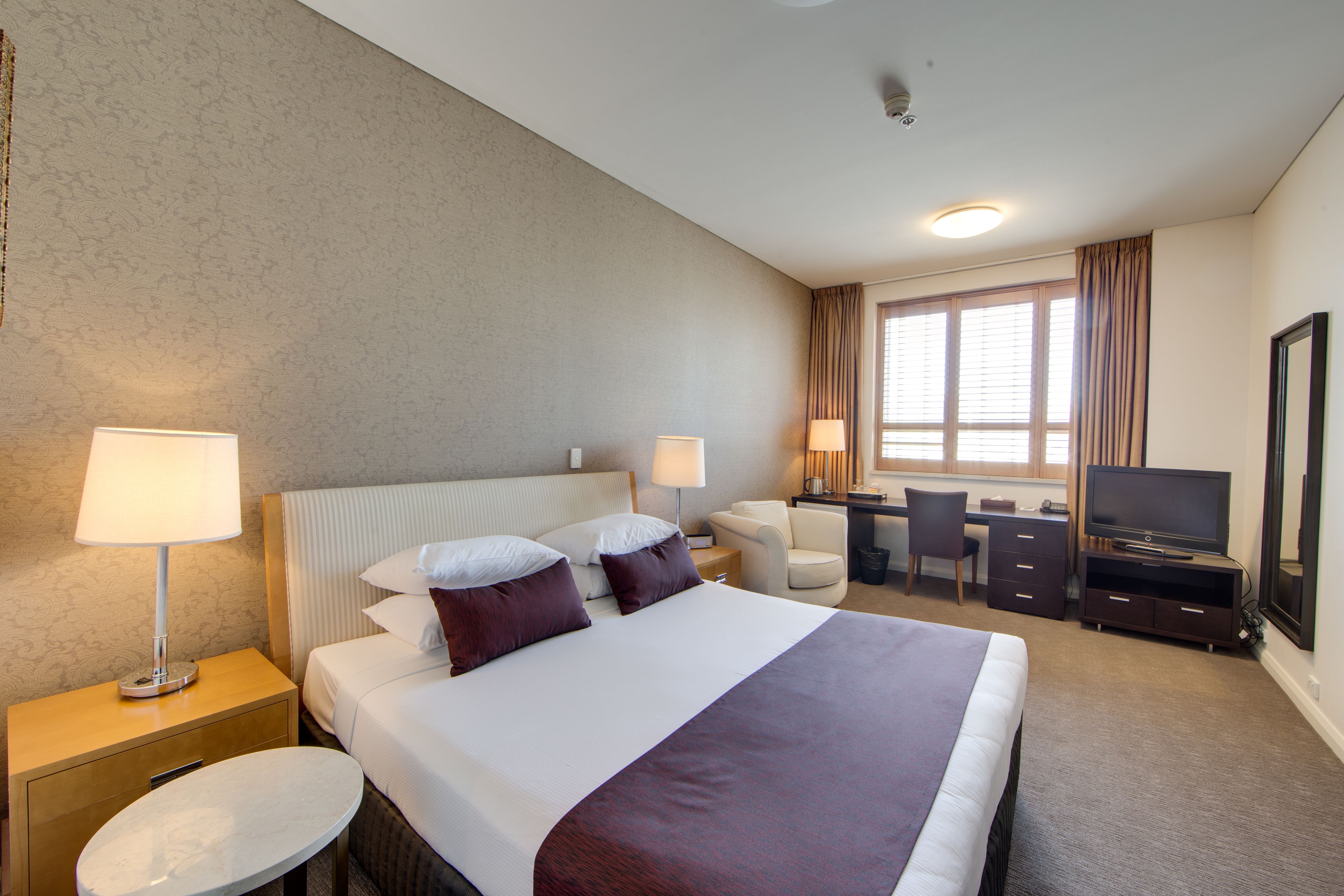 Habitación de hotel con una cama grande, dos lámparas de noche, un escritorio con silla, un televisor y un ventanal con cortinas.