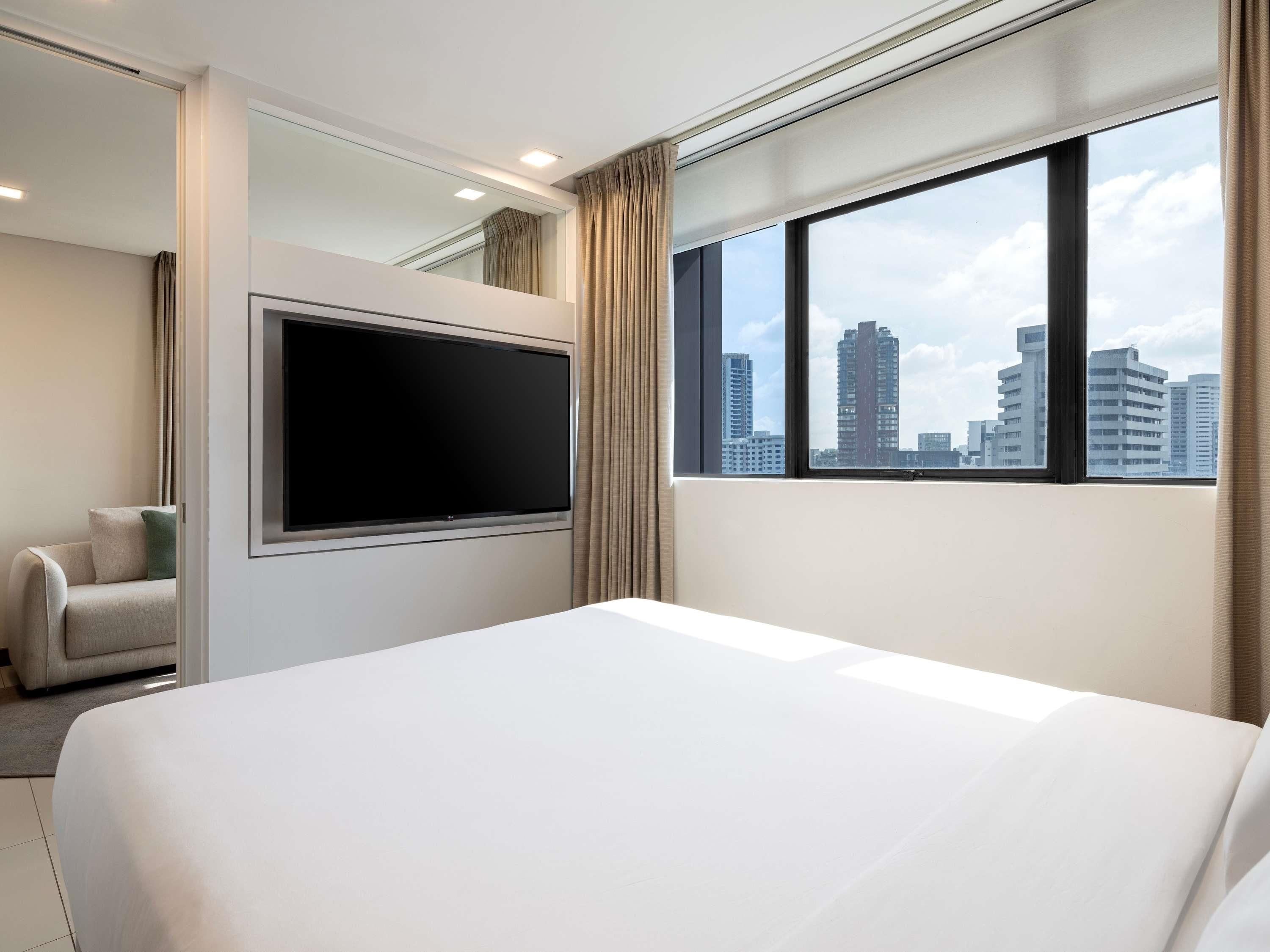 Une chambre d’hôtel dotée d’un grand lit, d’un téléviseur à écran plat fixé au mur, offrant une vue sur la ville grâce à une grande fenêtre, et comprenant un coin salon avec un fauteuil.