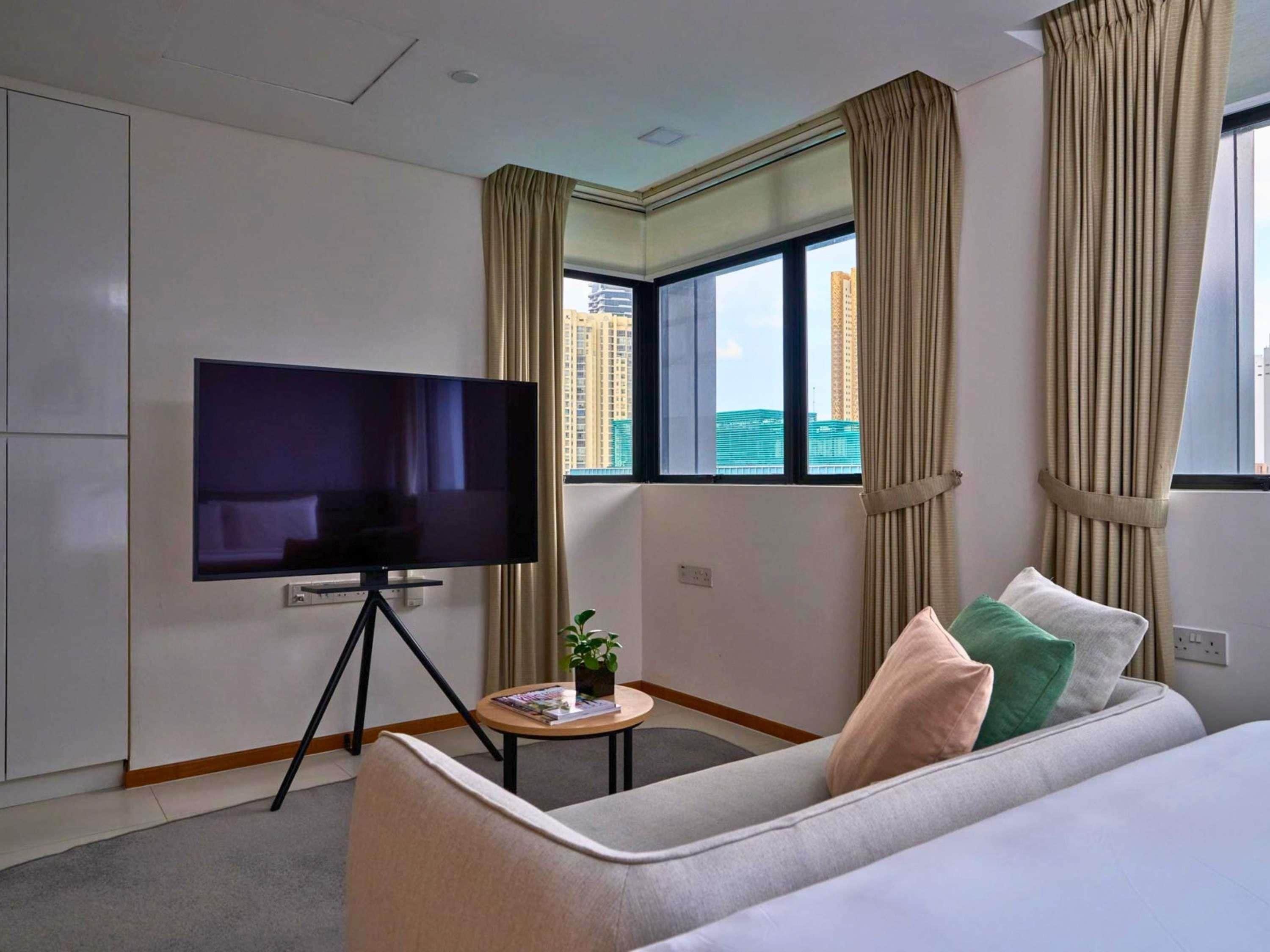 Une chambre d’hôtel équipée d’un téléviseur à écran plat sur un trépied, d’un canapé avec des coussins, d’une petite table avec une plante et d’une fenêtre avec des rideaux.