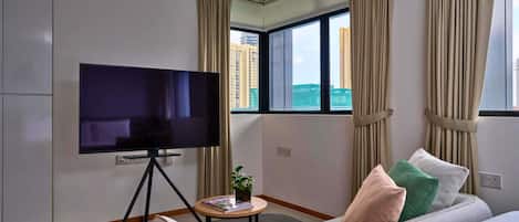Une chambre d’hôtel équipée d’un téléviseur à écran plat sur un trépied, d’un canapé avec des coussins, d’une petite table avec une plante et d’une fenêtre avec des rideaux.