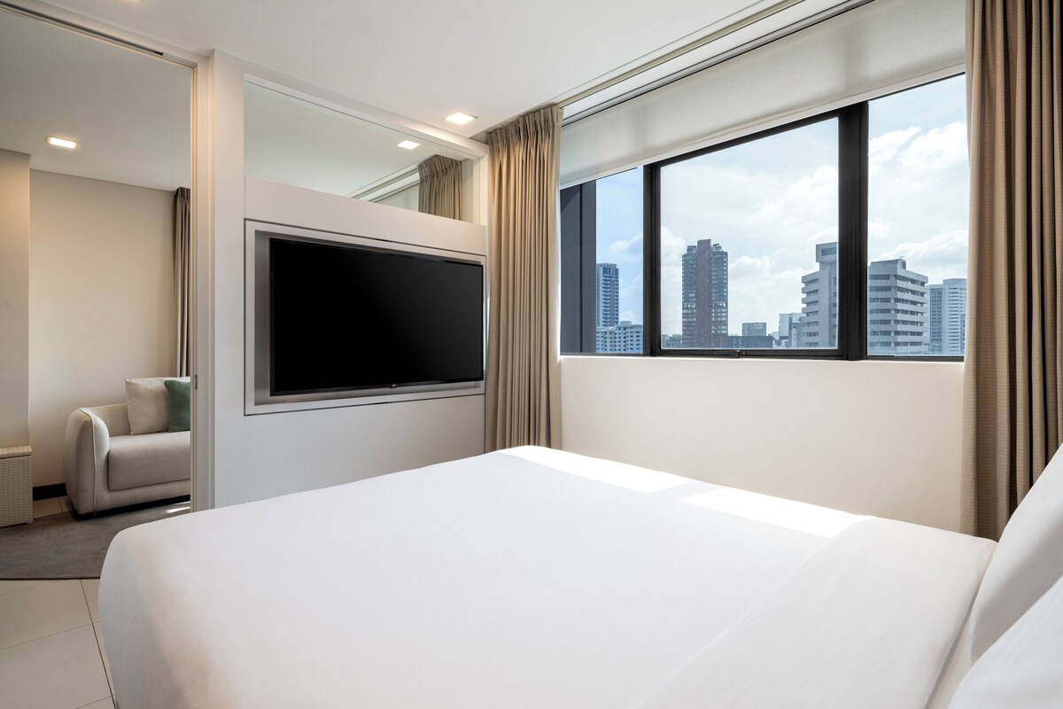 A hotel room with a large bed, a flat-screen TV, a sofa, and a city view.
