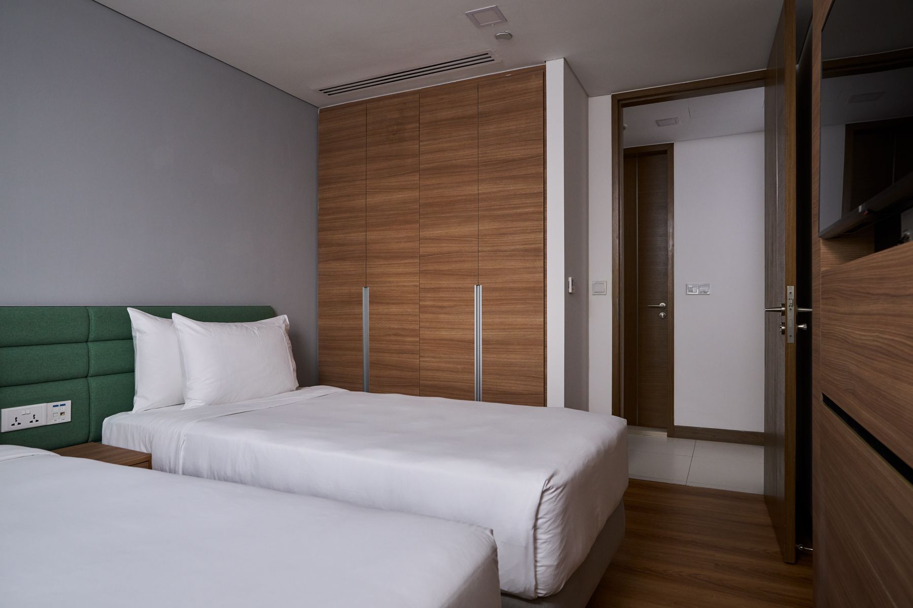 Un dormitorio con dos camas, un cabecero verde, un armario de madera y una puerta que da a otra habitación.