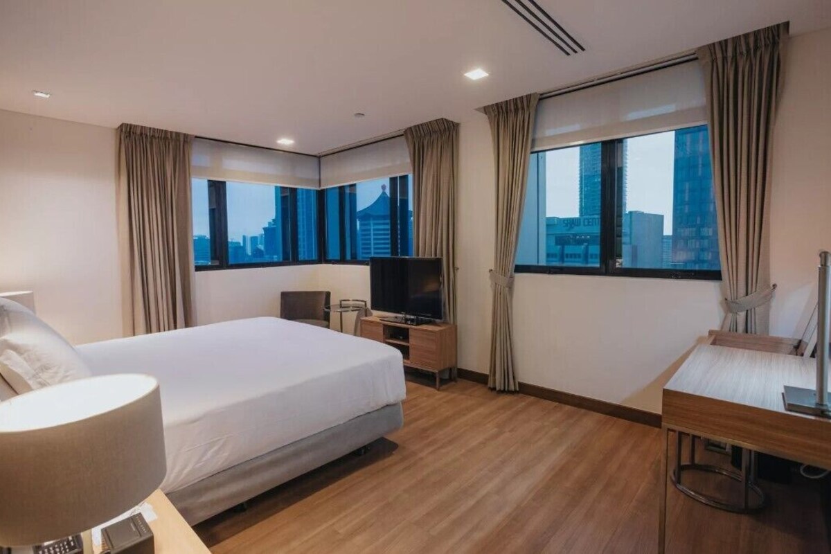 A hotel room with a large bed, a desk with a chair, a TV, and a view of the cityscape through large windows.