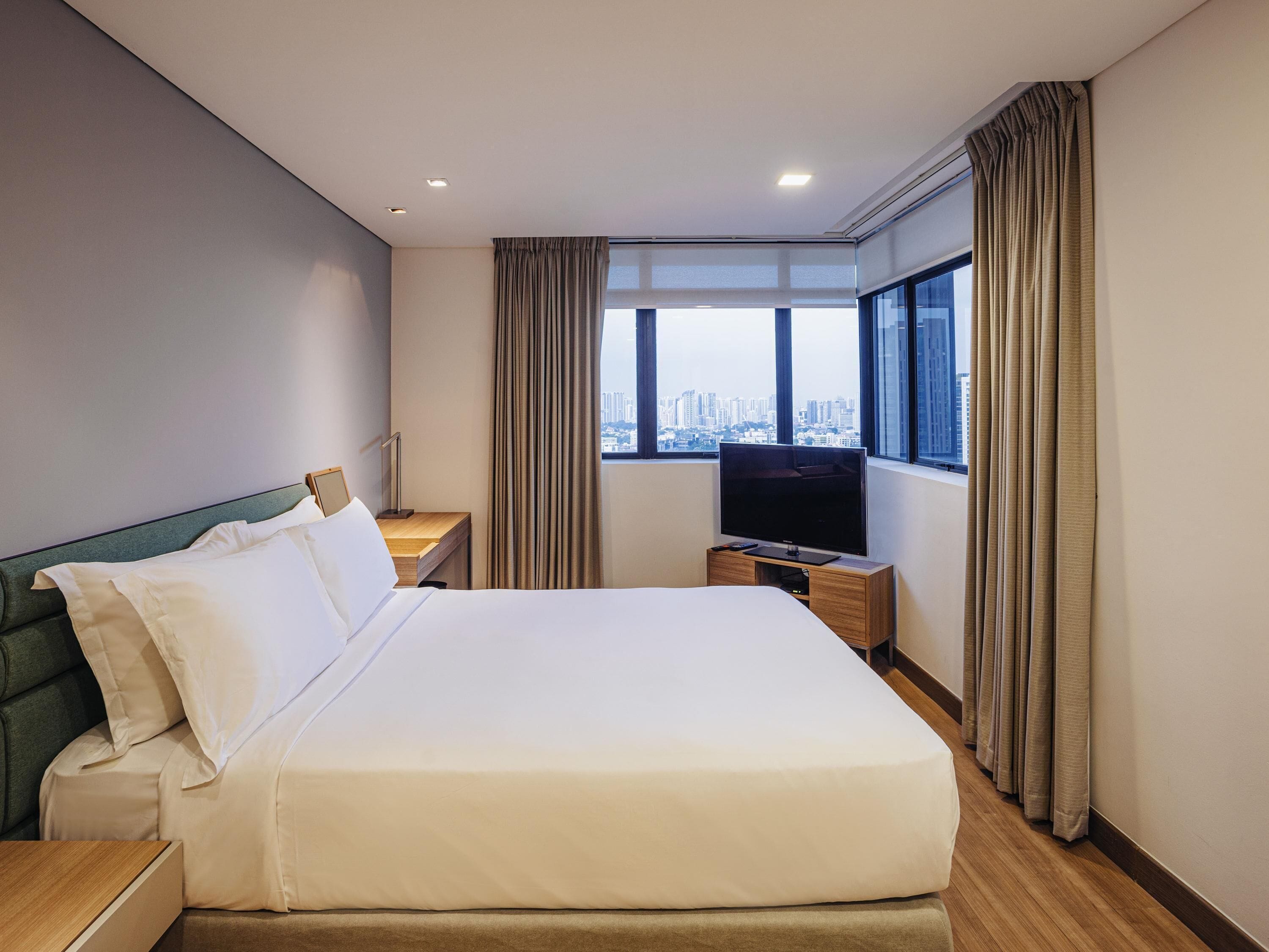 Ein Hotelzimmer mit einem großen Bett, einem Fernseher, einem Schreibtisch und einem Fenster mit Blick auf die Stadt.