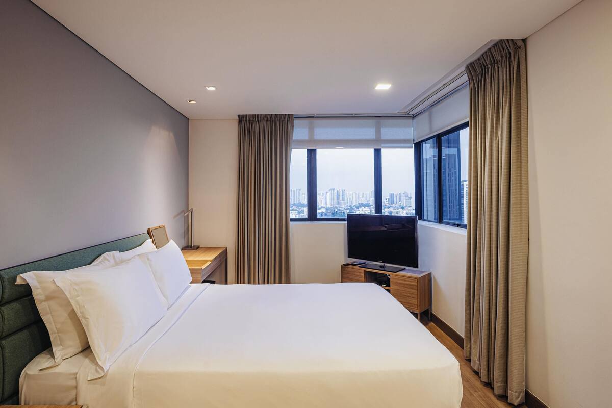 A hotel room with a large bed, a TV, a desk, and a window with a city view.