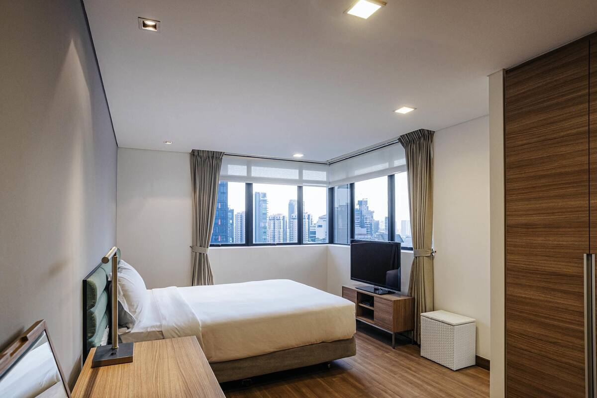 A modern hotel room with a bed, a desk, a TV, and a view of the cityscape.