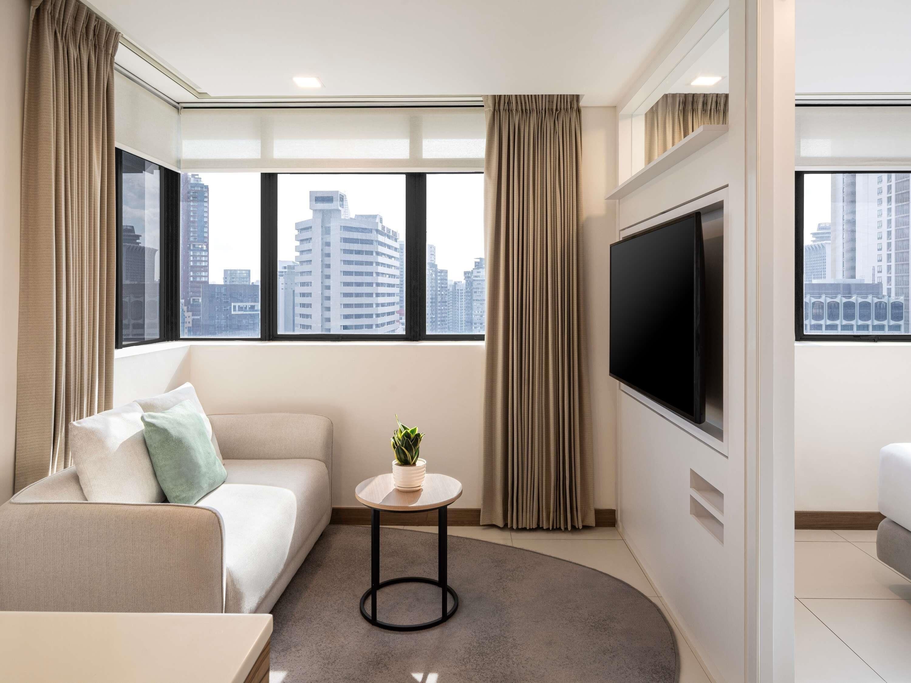 Ein modernes Hotelzimmer mit einer Couch, einem kleinen Tisch mit Pflanze, einem Fernseher und großen Fenstern mit Blick auf die Stadt.