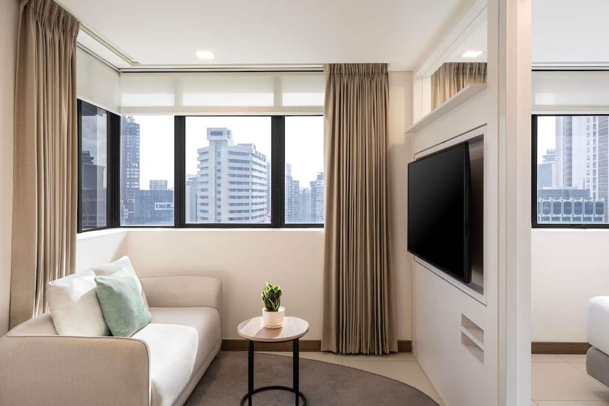 Ein modernes Hotelzimmer mit einer Couch, einem kleinen Tisch mit Pflanze, einem Fernseher und großen Fenstern mit Blick auf die Stadt.