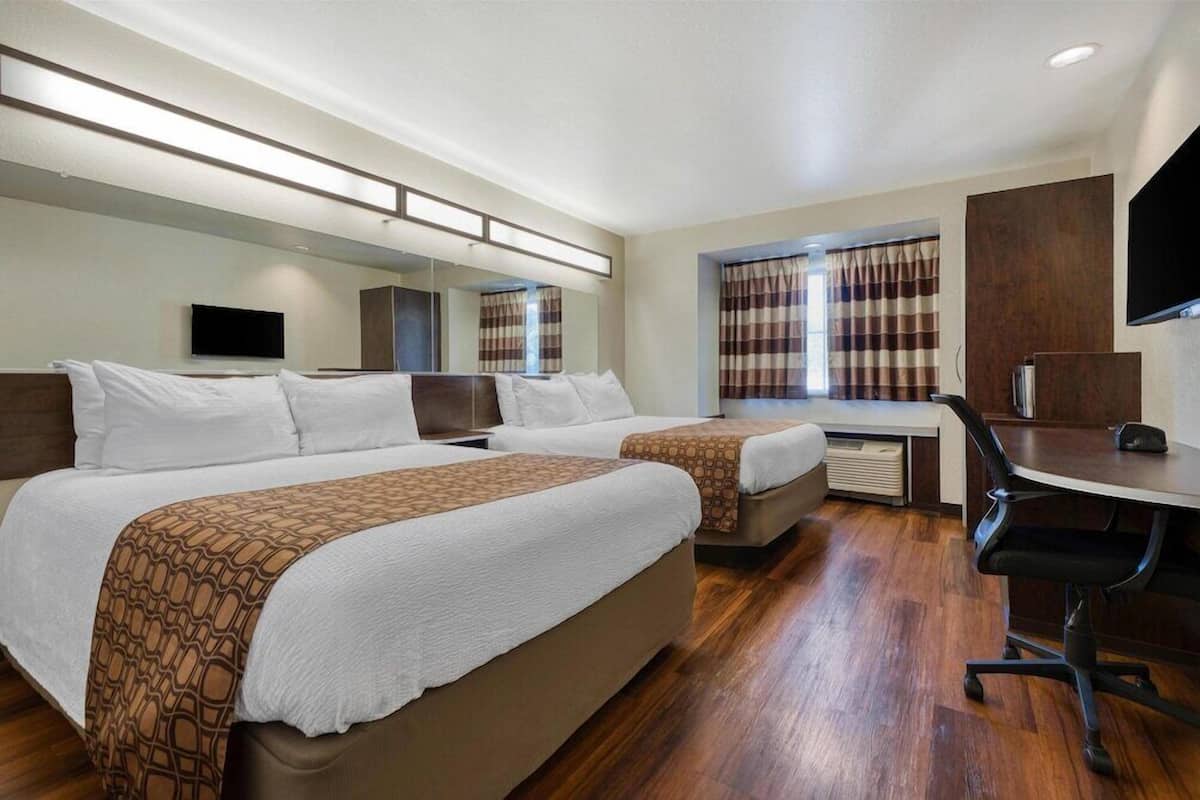 Habitación de hotel con dos camas, un escritorio, una silla, un televisor de pantalla plana y un ventanal grande con cortinas.