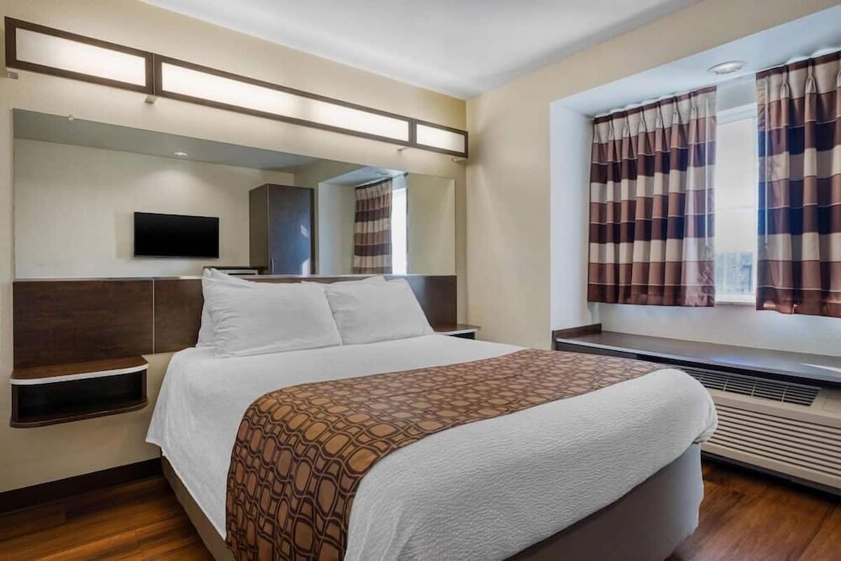Una habitación de hotel con cama, mesitas de noche, televisor de pantalla plana, ventana con cortinas y espejo montado en la pared.
