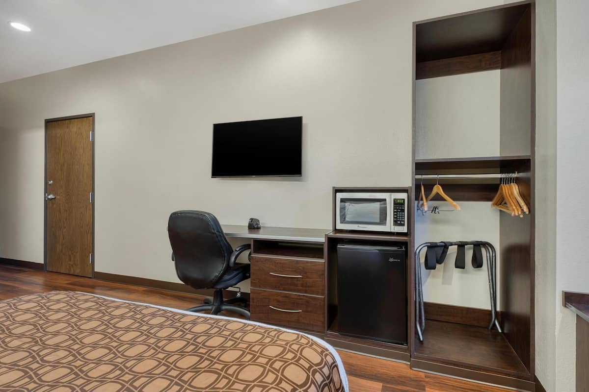 Habitación de hotel con cama, escritorio con computadora, microondas, refrigerador pequeño, televisor y armario con ropa colgada.