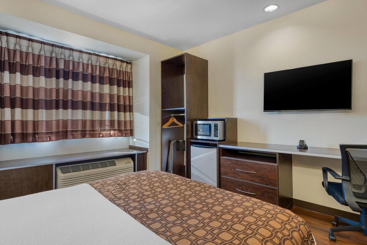 Habitación de hotel con cama, escritorio con computadora, televisión, microondas y refrigerador pequeño.