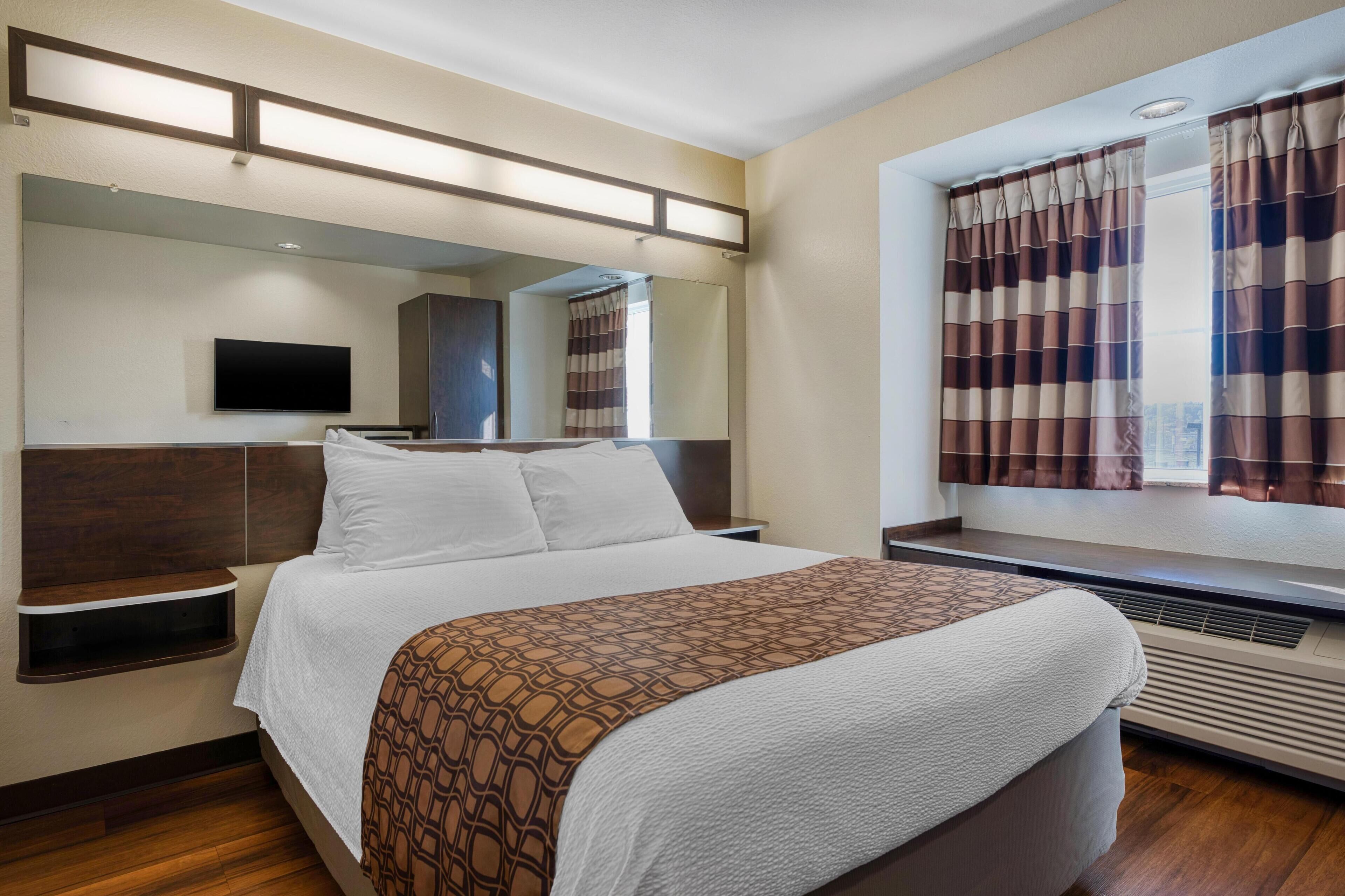 Ein Hotelzimmer mit einem großen Bett, einem Flachbildfernseher, einem Fenster mit Vorhängen und einer fest installierten Heizung.