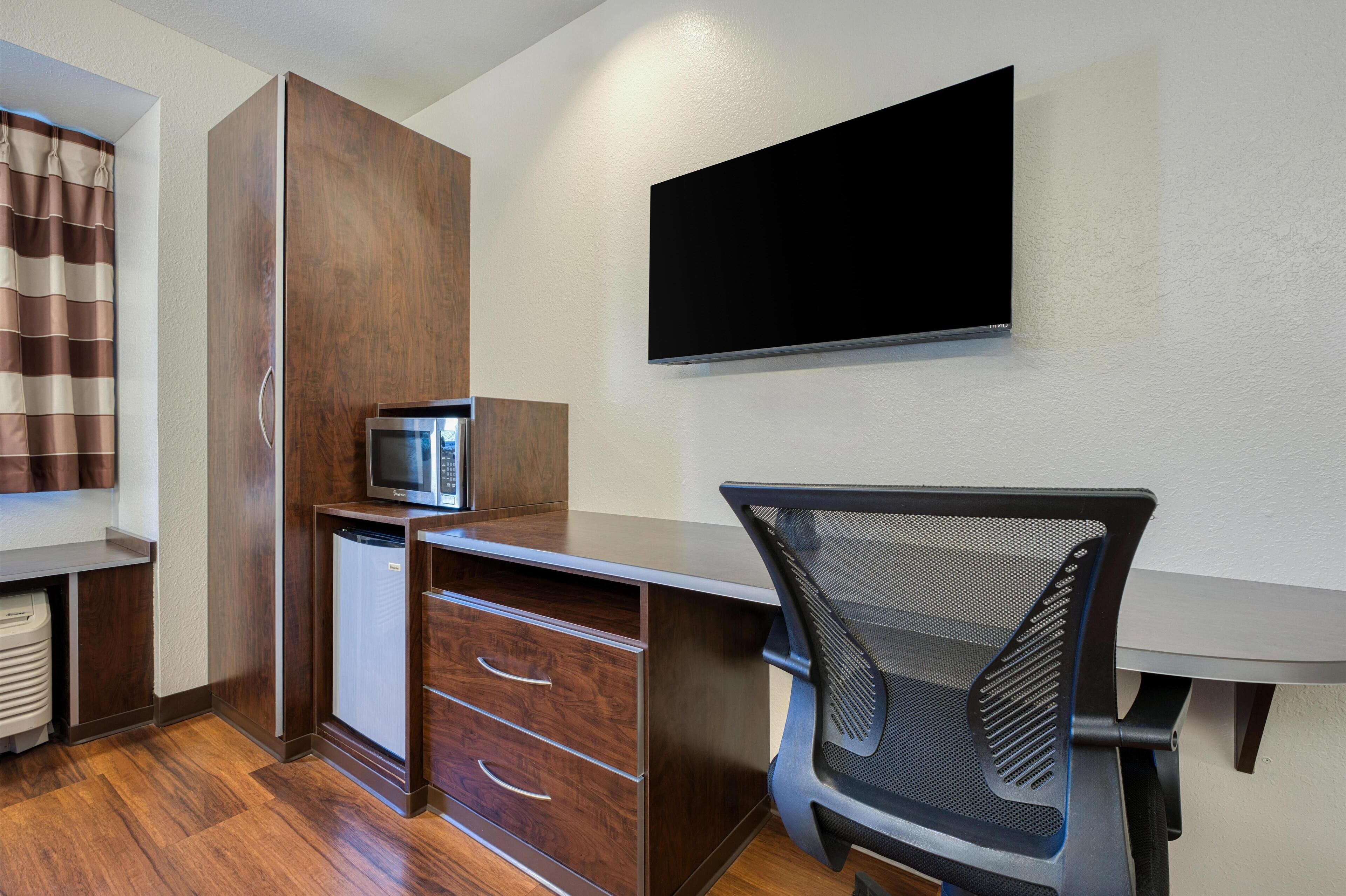 A compact hotel room with a wooden desk, a flat-screen TV, a small refrigerator, and a foldable chair.
