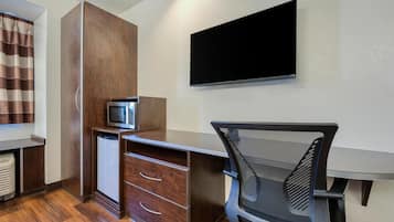 A compact hotel room with a wooden desk, a flat-screen TV, a small refrigerator, and a foldable chair.