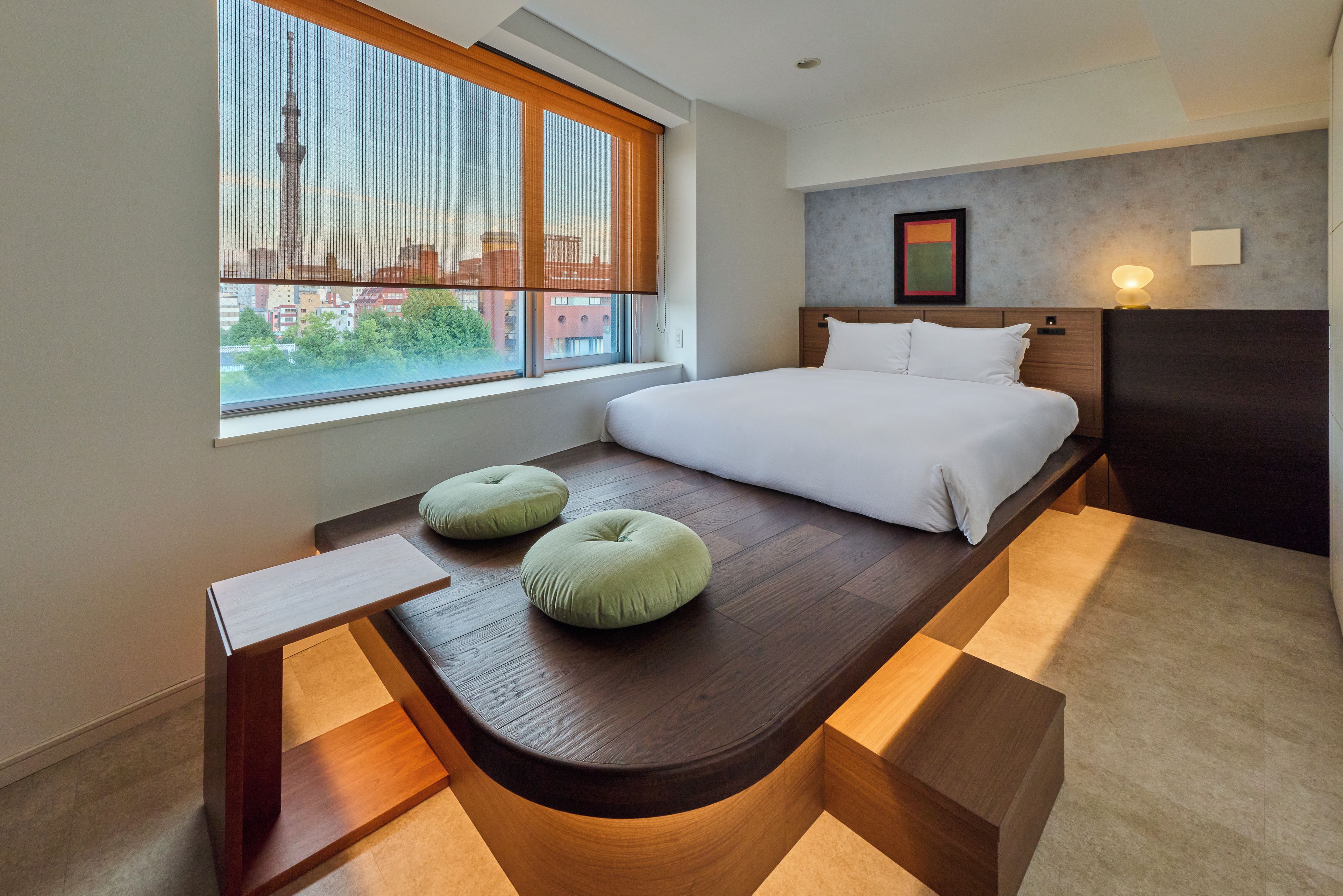 Una habitación de hotel moderna con una cama grande, un banco con cojines, una ventana con vistas a edificios y una lámpara de pared.