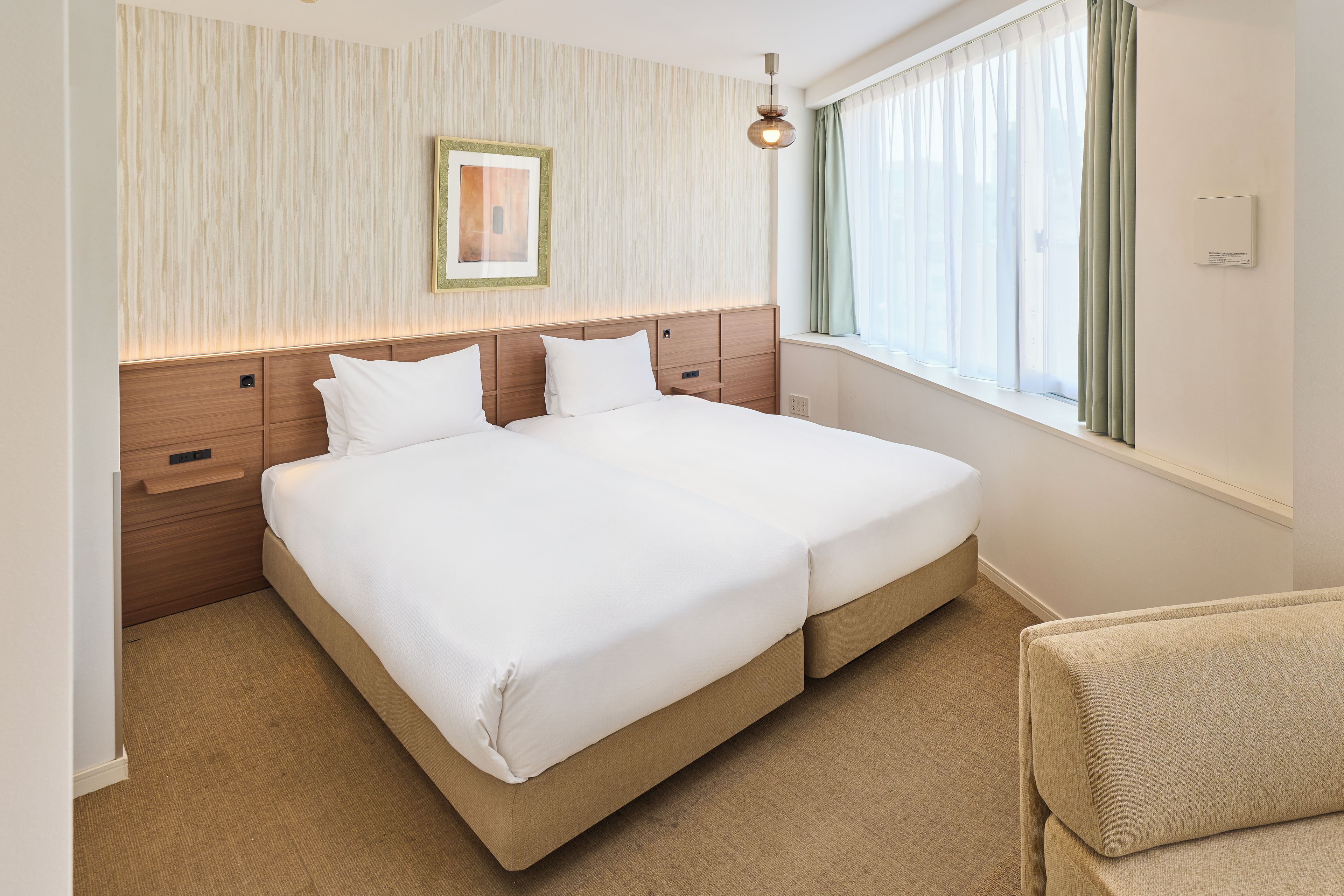 A hotel room with a double bed, wooden headboard, a bedside table, a small bedside lamp, a window with curtains, and a wall-mounted picture frame.
