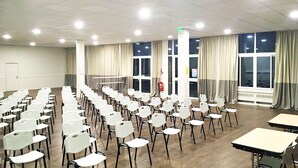 A conference room with rows of white chairs and tables, large windows with curtains, and a fire extinguisher on the wall. - Complexe Hôtelier Regain (Sainte-Tulle)