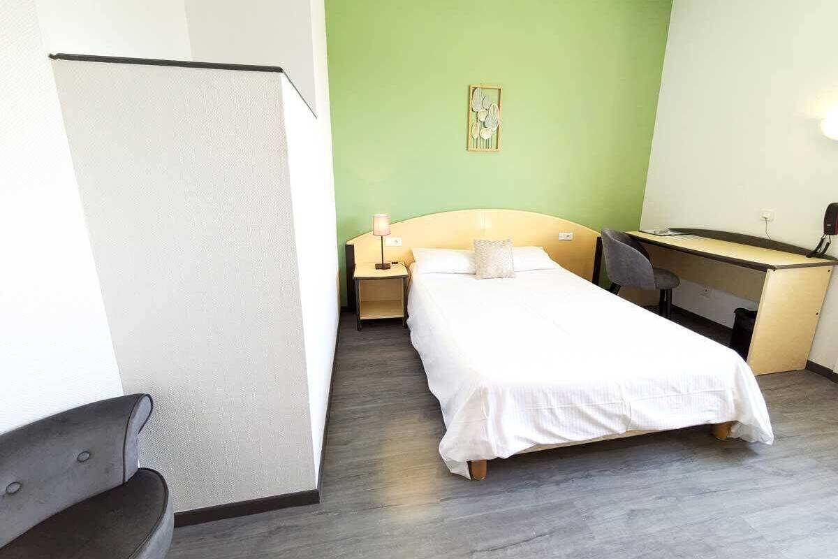 A hotel room with a bed, a desk, a chair, a nightstand, and a wall-mounted lamp.