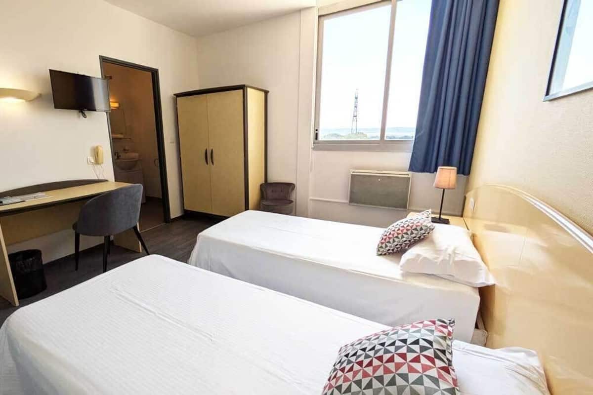 A hotel room with two beds, a desk, a chair, a wardrobe, a TV, and a window with curtains.