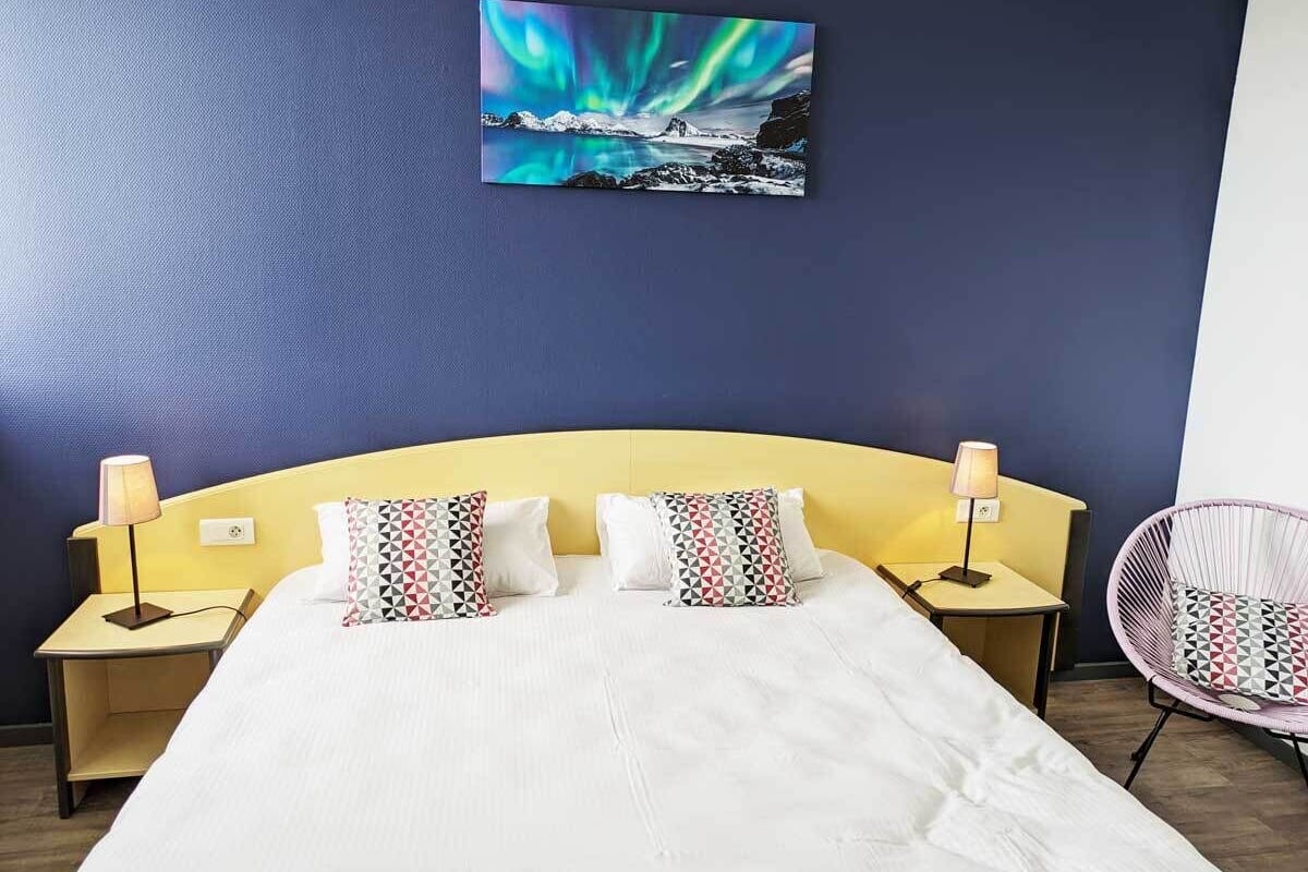 A hotel room with a bed, two bedside tables with lamps, a chair, and a painting of the Northern Lights on the wall.