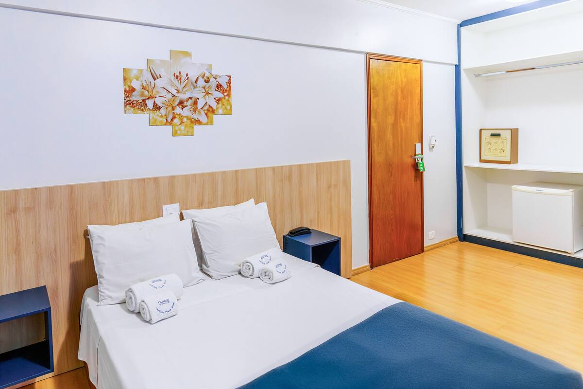 Une chambre d’hôtel avec une tête de lit en bois, un lit recouvert de draps blancs, une couverture bleue, une table de chevet avec un téléphone et une œuvre d’art accrochée au mur.