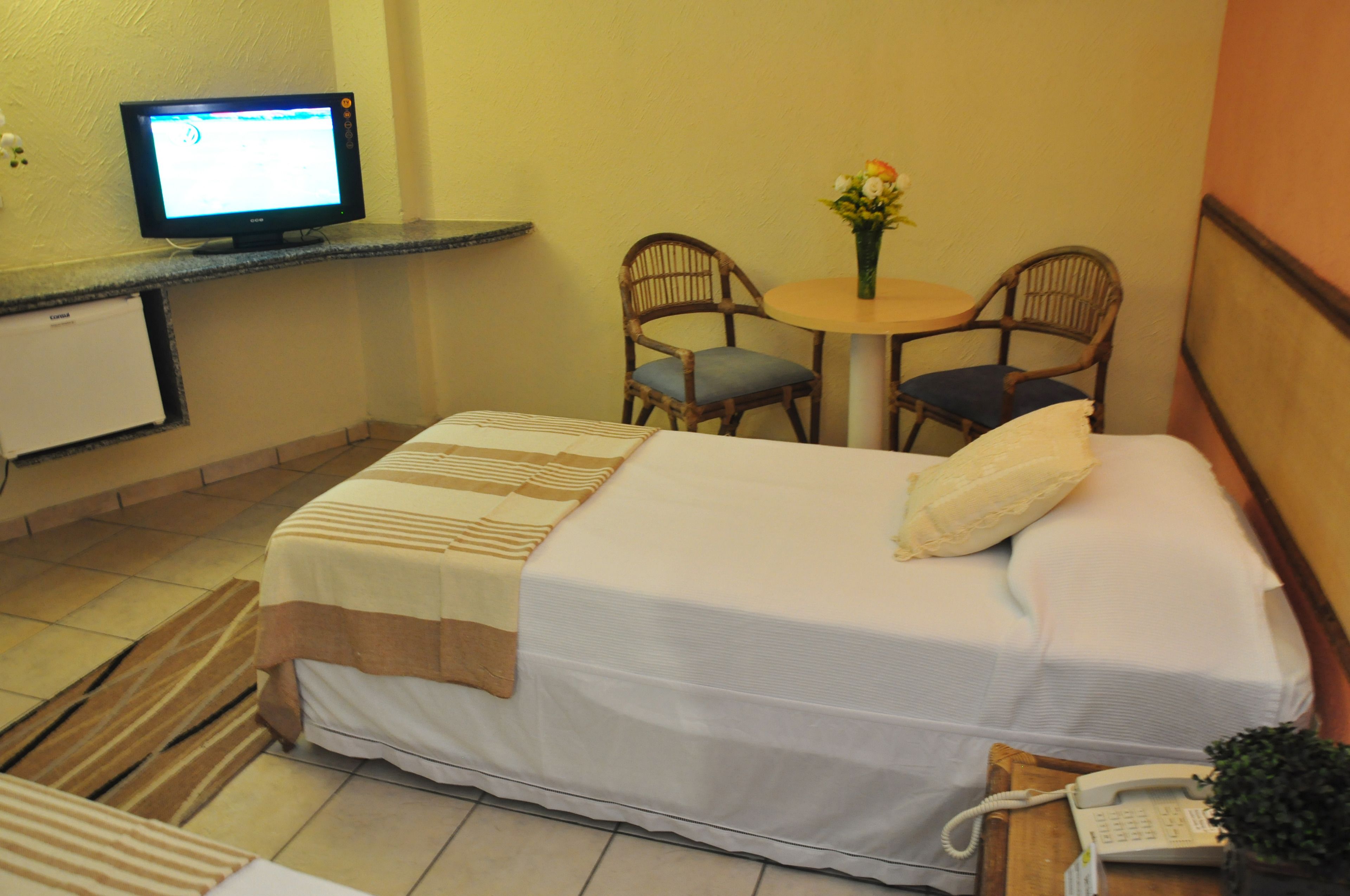 Une chambre d’hôtel comprenant un lit, un bureau avec un ordinateur, deux chaises, une petite table avec un vase et un téléphone sur une table d’appoint.
