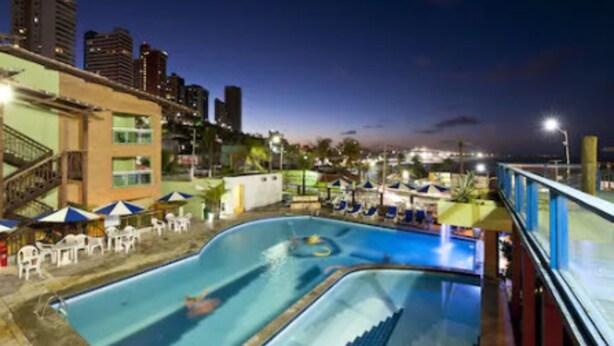 A swimming pool with lounge chairs and umbrellas, surrounded by buildings and a city skyline at dusk. - Natal Praia Hotel (Natal)