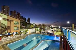 Piscina com espreguiçadeiras e guarda-sóis, cercada por edifícios e um horizonte urbano ao entardecer.