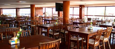 A spacious dining area with multiple wooden tables and chairs, large windows, and a television mounted on the wall.