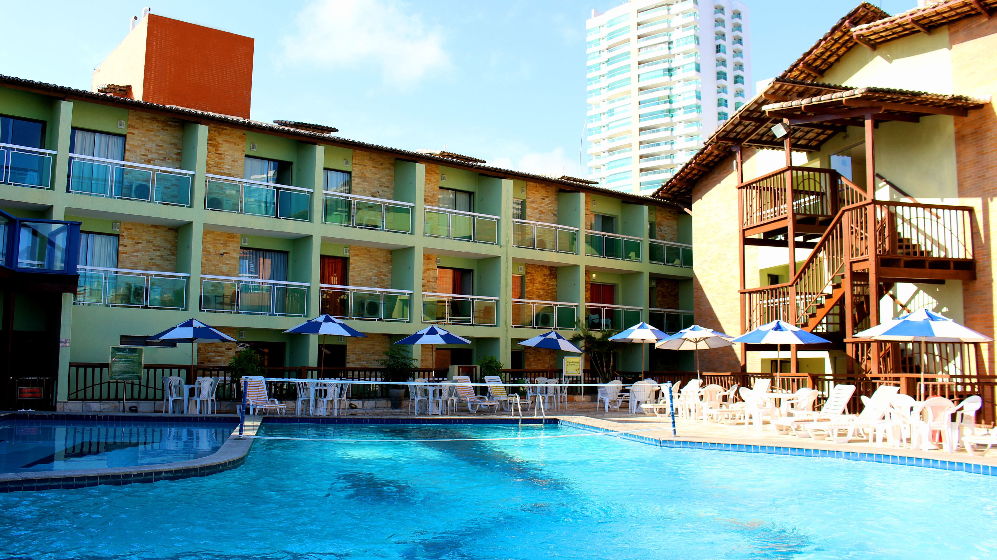 A hotel pool area with multiple lounge chairs and umbrellas, a swimming pool, and a building with balconies and a staircase.