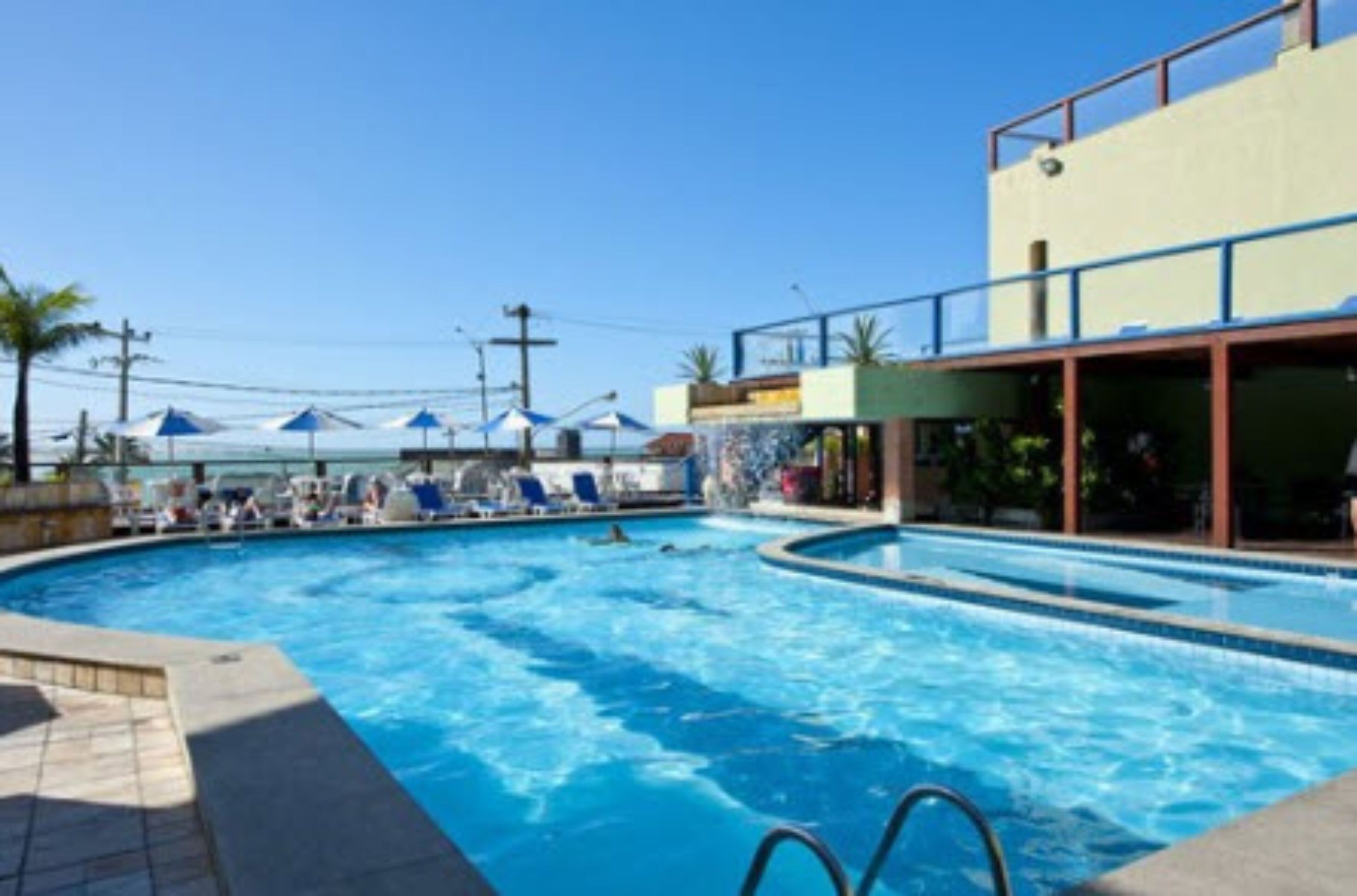 A swimming pool with lounge chairs and umbrellas, surrounded by a building with a balcony.