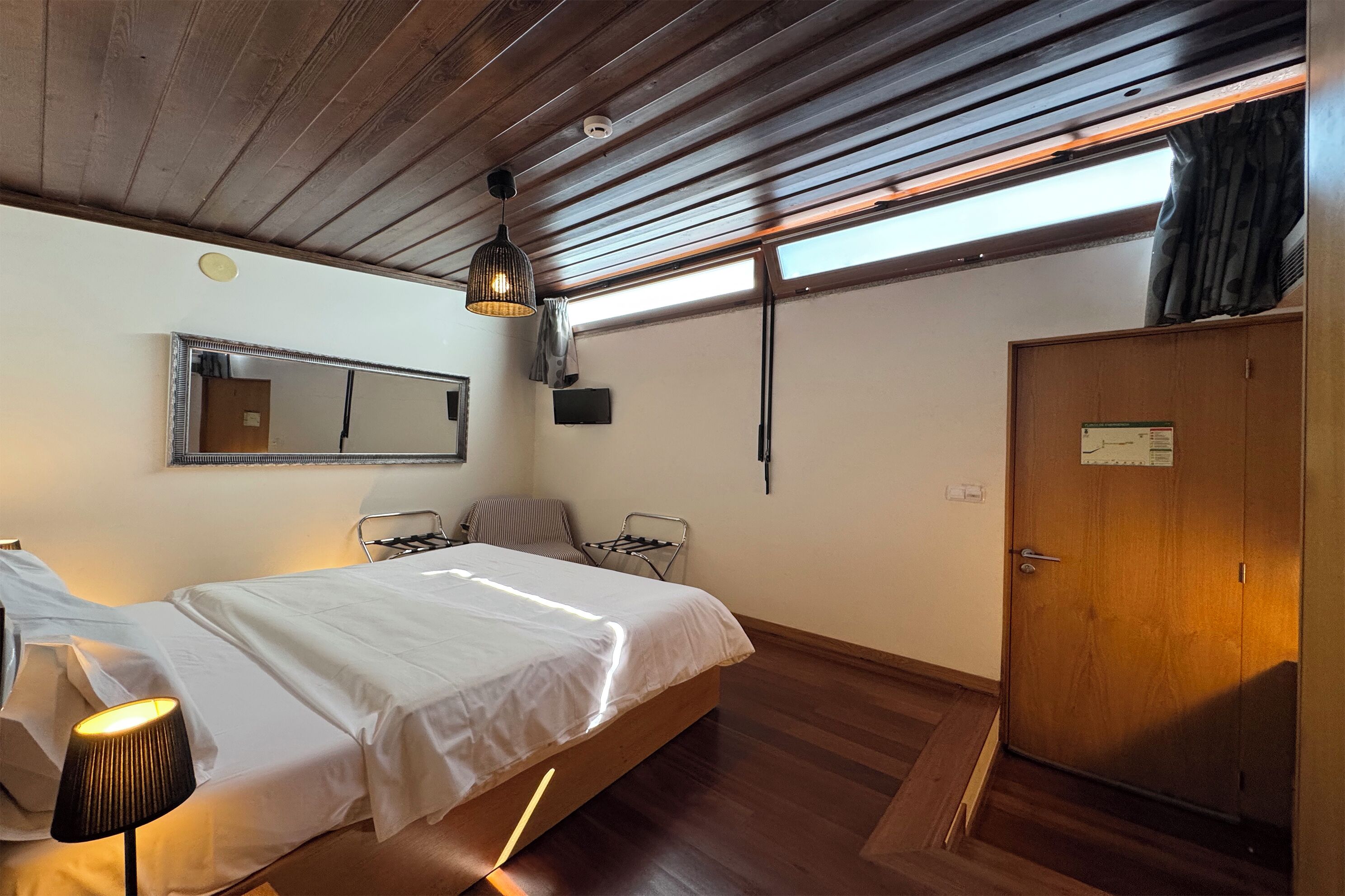 Um quarto com cama, uma cadeira, um guarda-roupa, um espelho e um candeeiro no teto.